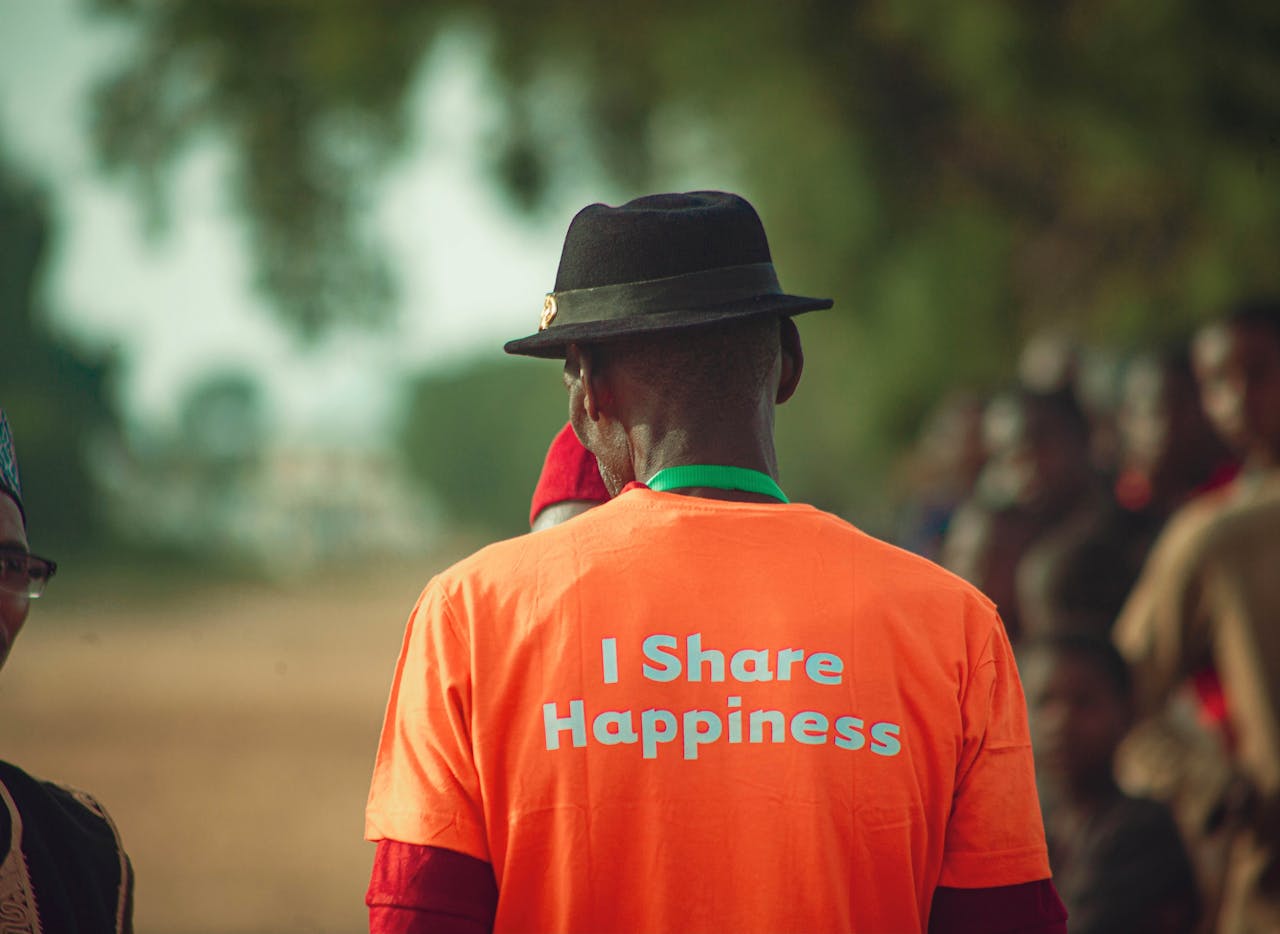 gallery-6 A man in an orange shirt with a happiness message stands outdoors, fostering positivity and community spirit.