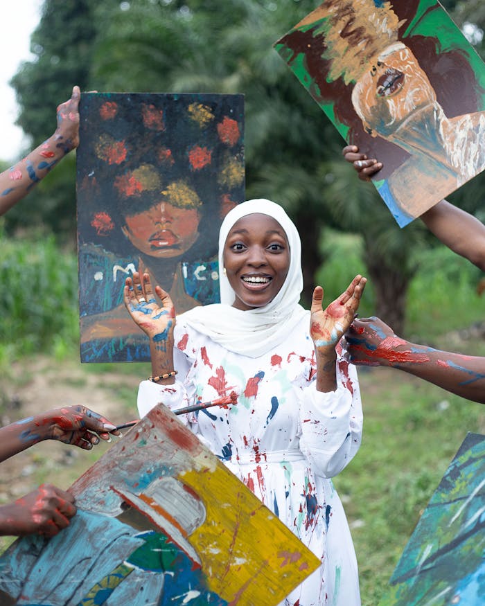 about-img Young artist in white hijab with vibrant paintings, smiling and joyful, outdoor setting.