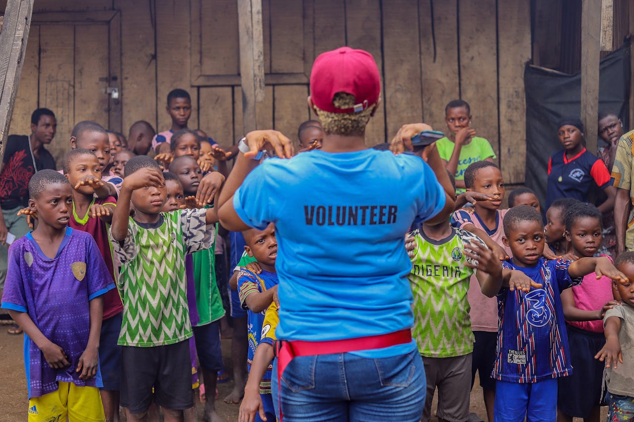 contact-img A volunteer guides a diverse group of children in an outdoor activity, fostering community spirit and teamwork.