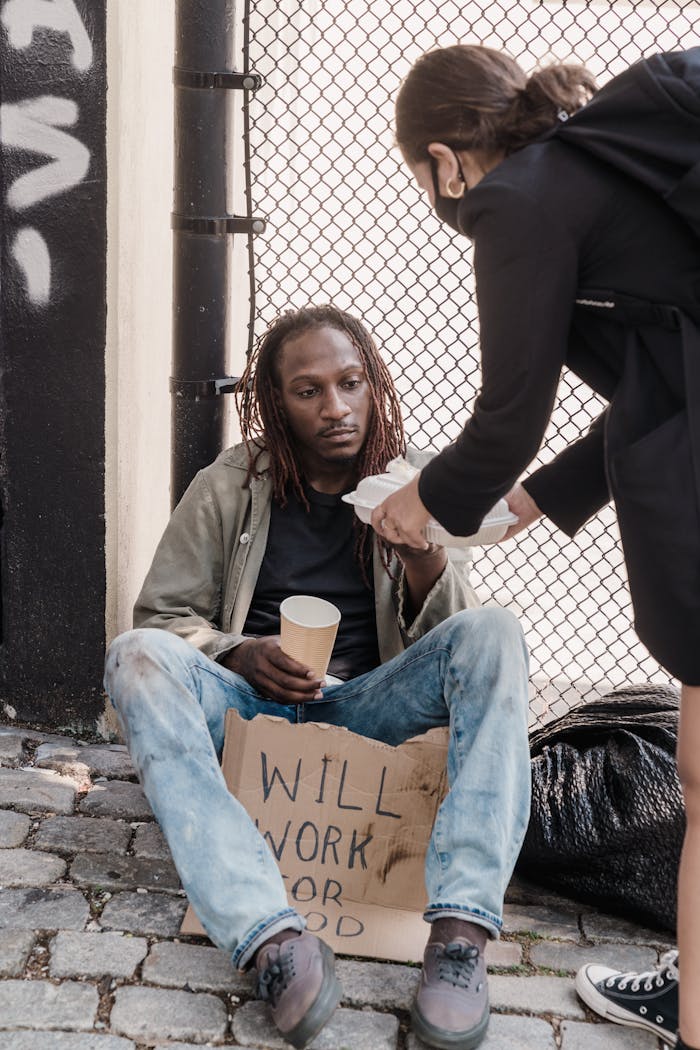 gallery-1 A woman offers food to a man sitting on the sidewalk holding a sign, showcasing empathy.