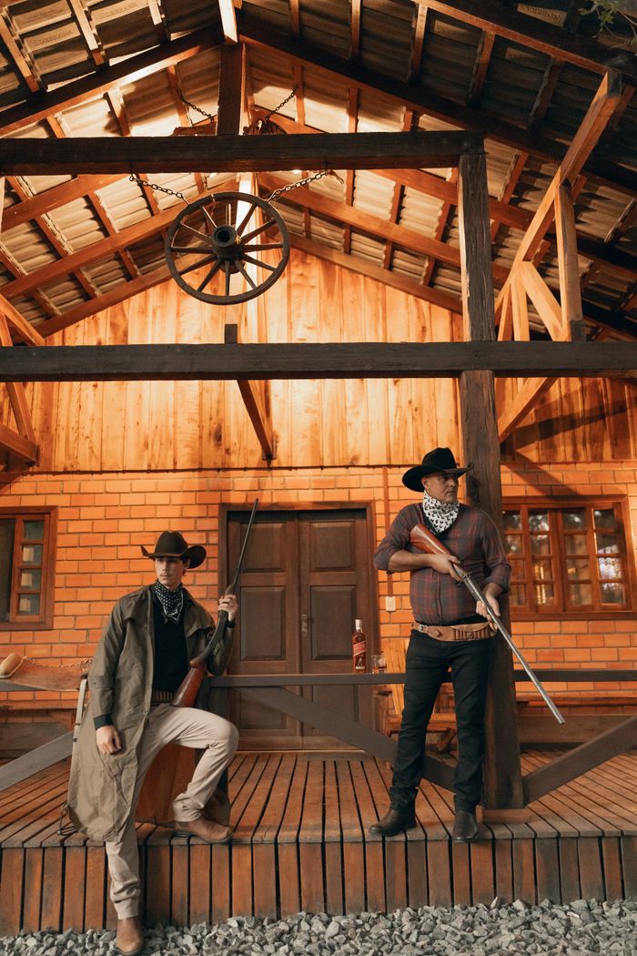 journey-01 Two cowboys with bandanas and rifles stand guard at a rustic cabin, evoking a classic Western scene.
